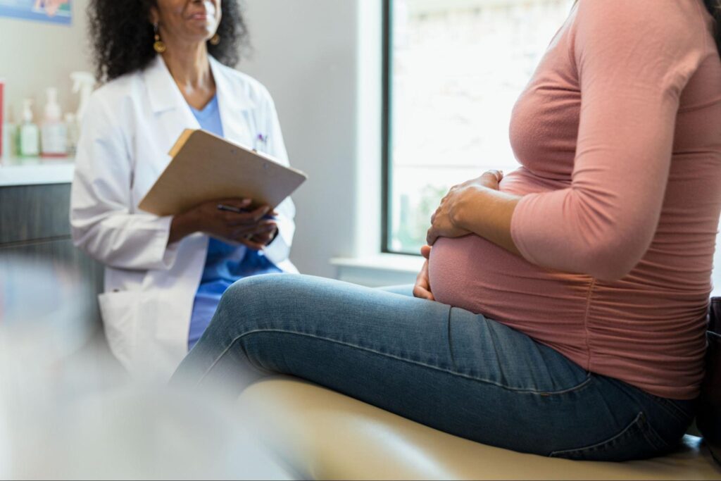 MOGA OB/GYN holding a clipboard and reviewing notes with a pregnant patient in a pink top who rests her hand on her belly during a prenatal appointment