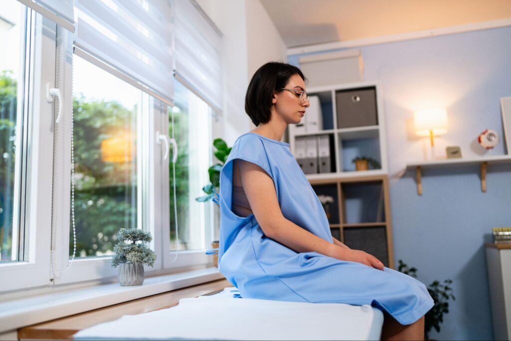 Patient in a light blue exam gown sitting on an exam table by a bright window, waiting for her prenatal appointment to begin at MOGA