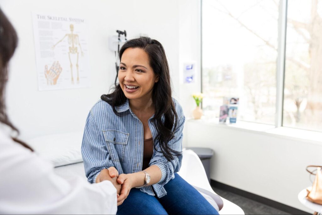 Smiling woman in a denim shirt sits on an exam table at MOGA, relaxed and waiting for her gynecologist appointment in a Memphis OB/GYN office
