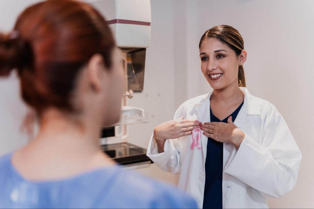 MOGA OB/GYN in a white coat holds a pink breast cancer awareness ribbon while talking with a patient next to a mammography machine, showing the preventive screenings a gynecologist offers